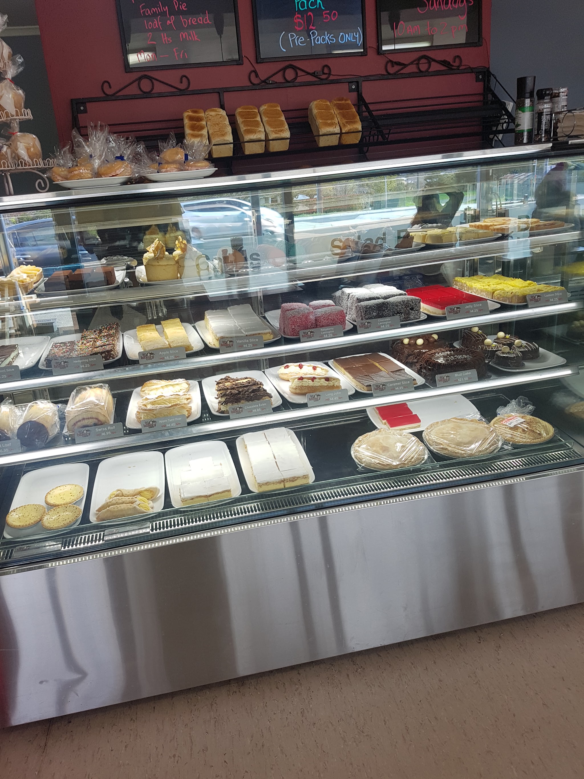 Cake cabinet full of slices, lamingtons and cakes, with fresh loaves on the shelf above